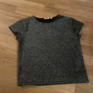 Glittrig svart topp från KappAhl - Snygg svart glittrig topp från KappAhl med korta ärmar och rund halsringning. Perfekt när du vill sticka ut lite extra. Toppens material har en skimrande yta som fångar ljuset och ger en cool look.