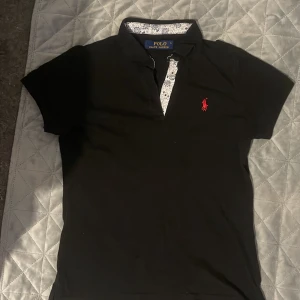 Svart skjorta från Polo Ralph Lauren - Svart skjorta från Polo Ralph Lauren med korta ärmar och klassisk krage. Tröjan har knäppning med blommigt mönster på insidan och en röd broderad logga på bröstet. Tillverkad i mjuk bomull för en skön känsla. Perfekt för en clean och stilren look. Knappast använt!!