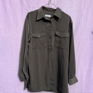 Mörkgrå skjort från Friendtex - Stilren mörkgrå skjorta från Friendtex i polyester. Skjortan har klassisk krage, två stora bröstfickor med lock och knappar framtill. Långärmad modell med breda manschetter och rak passform. Perfekt att bära öppen eller stängd för en avslappnad look.