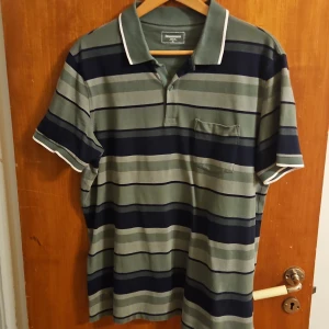 Randig pikétröja från Dressmann XL - Snygg randig pikétröja från Dressmann i grönt och marinblått med klassisk krage och knappslå. T-shirten har korta ärmar, bröstficka och vita detaljer på krage och ärmslut. Perfekt för en chill och stilren look.