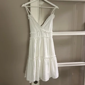 Vit omlottklänning - Superfin vit kortklänning med omlottliv, smala axelband och volangdetaljer. Klänningen har lätt struktur i tyget och är perfekt för sommarens alla tillfällen. Söt och luftig modell med markerad midja och volanger i flera lager. Korsade band och dragkedja i ryggen