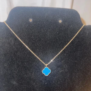 Guldhalsband med blå fyrklöverberlock - Snyggt halsband i guld med en blå fyrklöverformad berlock. Kedjan är tunn och elegant, perfekt för att ge din outfit en färgklick. Berlocken har en mjuk, rundad form och är infattad i guldfärgad metall. Passar dig som gillar stilrena smycken med lite extra edge.