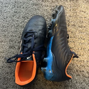 Adidas copa fotbollsskor - Säljer ett par Adidas copa i mörkblått skinn med orange innersida och detaljer. Skorna har klassiska vita Adidas-ränder på sidan och blå sula med dobbar för gräsplan. Perfekta för dig som vill sticka ut på planen. Skorna är använda i cirka ett halvår men funkar för dig som vill ha ett par prisvärda skor. Storlek 36.