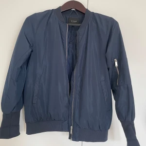 Mörkblå bomberjacka från Vila - Snygg mörkblå bomberjacka från Vila med klassisk ribbad mudd vid ärmslut och midja. Jackan har dragkedja framtill och en extra ficka med dragkedja på ena ärmen. Perfekt för dig som gillar en stilren och enkel look.