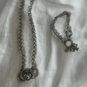 Guess halsband och armband - Säljer ett halsband och matchande armband från Guess i silverfärgad metall. Halsbandet har tre ringar, varav en med Guess-logga, och armbandet har berlocker med bokstäverna GUES samt en glittrig S. Båda har grova länkar och coola detaljer