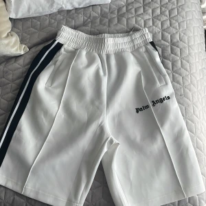 Vita Palm Angels shorts med svarta ränder - Palm Angels vita shorts med svarta sidoränder och broderad logga på vänster ben. De har elastisk midja med snörning och är tillverkade i ett slätt, sportigt polyestermaterial. Perfekta för en clean och sportig look.