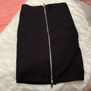 Svart kortkjol med dragkedja - Snygg svart kortkjol med en silverfärgad dragkedja som går hela vägen framtill. Kjolen har en rak och tight passform som ger en cool och stilren look. Materialet känns mjukt och stretchigt, perfekt för att skapa en edgy outfit.