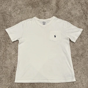 Vit v-ringad t-shirt Polo Ralph Lauren - Vit t-shirt från Polo Ralph Lauren med v-ringning och korta ärmar. På bröstet finns en ficka med den klassiska broderade Polo-loggan i mörkblått. Tillverkad i mjuk bomull för skön känsla och clean look.