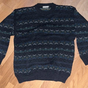 Mörkgrön stickad  - Säljer en mörkgrå stickad tröja från Avenue med snyggt mönster i blått, grönt och gult. Tröjan har rund hals och långa ärmar, perfekt för dig som gillar retrovibbar och vill ha något varmt och stilrent. Passar dig som gillar klassisk stil med en twist.