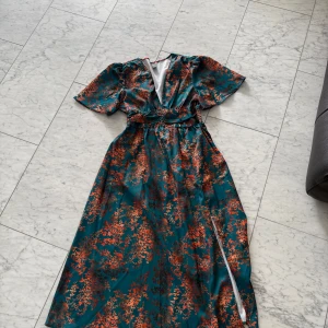 Blommig långklänning med slits - Säljer en snygg långklänning med blommigt mönster i orange och rött mot en mörkgrön bakgrund. Klänningen har korta ärmar, v-ringad hals och en slits på sidan. Midjan är markerad med ett band och tyget har en glansig satinliknande finish. Perfekt för dig som vill sticka ut.