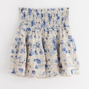 Blommig volangkjol från Lindex - Supersöt kort kjol från Lindex med blått blommönster på beige botten. Kjolen har bred smock i midjan, flera volanger och spetsdetaljer. Perfekt för vår och sommar när du vill ha en romantisk och luftig look.andvänd ca 1 gång