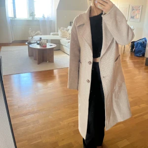 Beige kappa från KappAhl XS - Snygg beige kappa från KappAhl i storlek XS. Jackan har en klassisk krage, två knappar framtill och stora fickor. Materialet är mjukt och har en diskret rutig struktur som ger en stilren vibe. Perfekt för dig som gillar en clean och tidlös look.