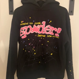Svart hoodie från Spider med stjärnor - Svart hoodie från Spider med färgglada stjärnor och ett stort tryck framtill med texten 'spider!'. Tröjan har huva, lång ärm och coolt spindelnätsmönster på magen. Perfekt för dig som gillar unika och lekfulla plagg. Materialet är mjuk bomullsmix. Kop1a såklart som alla sp5der tröjor