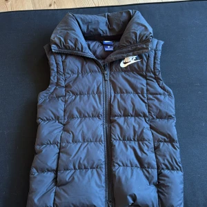 Svart dunväst från Nike - Snygg svart dunväst från Nike med hög krage och dragkedja framtill. Västen har quiltad design och ett vitt Nike-logga på bröstet. Perfekt att slänga över en hoodie för en sportig look. Materialet är syntet och dun, vilket gör den både lätt och varm.
