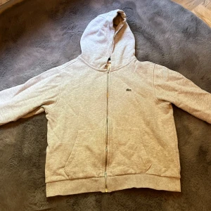 Grå hoodie från Lacoste med dragkedja - Snygg ljusgrå hoodie från Lacoste med dragkedja och huva. Klassisk krokodil-logga på bröstet och två fickor framtill. Mjuk insida och ribbade muddar. Perfekt för chill dagar eller när du vill ha en clean look. Det är storlek 164 men den då har krympt i tvätten. Den har inga missfärgningar och den ser nästan ut som ny. Jag köpte den för 1200.Priset kan diskuteras 