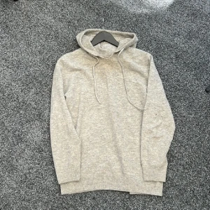 Ljusgrå stickad hoodie med huva - Mysig ljusgrå stickad hoodie med huva och snörning. Tröjan har långa ärmar och ribbade muddar vid ärmslut och nederkant. Perfekt för kyliga dagar när du vill vara både bekväm och stilren.trasig krage 