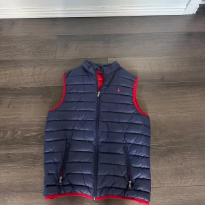 Mörkblå dunväst från Polo Ralph Lauren - Snygg mörkblå dunväst från Polo Ralph Lauren med röd insida och detaljer. Västen har horisontella quiltade sömmar, två dragkedjefickor och en diskret röd Polo-logga på bröstet. Perfekt för lager-på-lager. Storlek S men passar även XS. Pris ej hugget i sten, västen har knappast några tecken på användning👍 pris ej hugget i sten🙌