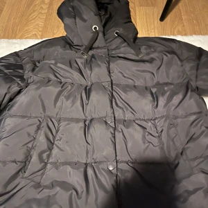 Svart pufferjacka med huva - Svart pufferjacka med huva och snörning, perfekt för kalla dagar. Jackan har långa ärmar, två sidofickor och knappar framtill. Materialet är syntet och jackan har en quiltad look som ger extra volym och värme. Enkel och stilren design som funkar till många outfits.