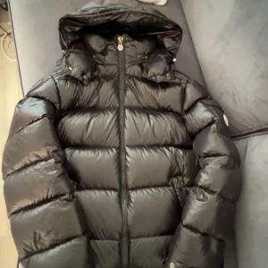 Moncler Maya - Svart pufferjacka från Moncler med glansig finish och huva. Jackan har breda, quiltade sektioner och dragkedja framtill. Insidan har en unik Moncler-serieetikett. Perfekt för dig som vill ha en snygg och varm jacka med streetkänsla. 