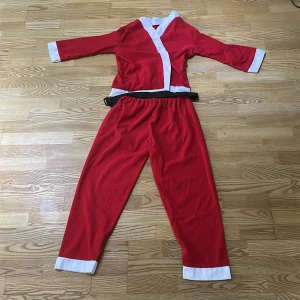 Röd tomtedräkt med bälte - Klassisk röd tomtedräkt med vita detaljer på ärmar, ben och krage. Dräkten består av en långärmad överdel och byxor med resår i midjan samt ett svart bälte. Perfekt för julens alla upptåg!