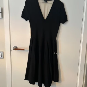 Svart kortklänning från Alexander McQueen - Svart kortklänning från Alexander McQueen med djup v-ringning, korta ärmar och en snygg detalj med metallring i midjan. Klänningen har en klassisk, lätt utställd siluett och är perfekt för dig som gillar stilren design med edge.