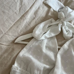 Vit kort klänning med rosett och volang - Supersöt vit kort klänning med broderade små blommor, omlottband som knyts i nacken och volang i kjolen. Smockad midja ger en snygg passform och tyget är lätt och luftigt, perfekt för varma dagar. Klänningen har en romantisk vibe och är riktigt fin på.