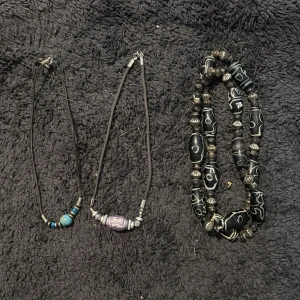 Svarta halsband med pärlor och mönster - Två coola halsband med svarta band och pärlor. Ett har blå och silverfärgade pärlor, det andra har en lila pärla med silverdetaljer. Det tredje halsbandet är gjort av svarta pärlor med vita mönster och metallpärlor. Perfekt för dig som gillar unik stil.