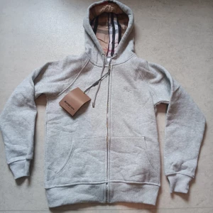 Grå hoodie från Burberry med dragkedja - Säljer en snygg grå hoodie från Burberry med klassiskt rutigt foder i huvan. Tröjan har dragkedja, snörning vid huvan och två stora fickor framtill. Perfekt för dig som gillar stilrena märkesplagg med exklusiv känsla och coola detaljer. Funkar till både sommar och vinter. 