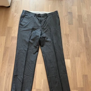 Grå kostymbyxor från Oscar Jacobson  - Snygga grå kostymbyxor från Oscar Jacobson, tillverkade i exklusivt tyg från Vitale Barberis Canonico. Byxorna har klassisk rak passform, sidfickor och är gjorda i en lyxig mix av 84% ull och 16% mohair. Perfekta för en stilren och dressad look.