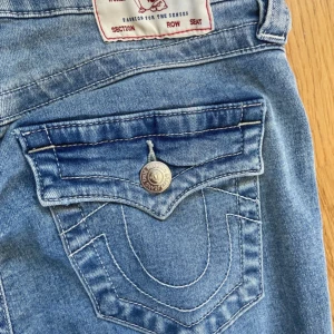 True Religion blå bootcut jeans - Snygga ljusblå jeans från True Religion med bootcut passform. Klassiska femficksmodellen med ikoniska broderade bakfickor och silverfärgade knappar. Jeansen har låg midja och är kanske använda 2 gånger och har absolut inga täcken på användning. Säljer pga att det inte passar längre  Storlek 26/32 kvittot finns kvar  Ordinarie pris 1 299