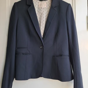 Mörkblå kavaj från Part Two, stl 36 - Snygg mörkblå kavaj från Part Two med klassisk krage och en knapp framtill. Fodret är ljust med prickigt mönster. Kavajen har två fickor med lock och är figursydd för en stilren look.