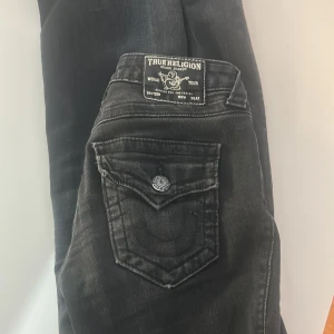 True Religion Becca Bootcut Jeans Svart - Svarta jeans från True Religion, modell Becca Mid Rise Bootcut. Snygga detaljer med markerade sömmar och ficklock bak med knappar. Klassisk bootcut-passform och cool tvättad look. Perfekta för dig som gillar street och premiumkänsla.