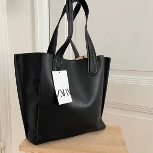 Svart shoppingväska från Zara - Stilren svart shoppingväska från Zara. Väskan har två rejäla handtag och är tillverkad i slätt syntetmaterial med lätt struktur. Perfekt för att bära allt du behöver till skolan eller stan. Enkel och klassisk design som funkar till det mesta.