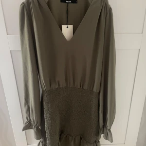 grön klänning med volanger - Supersnygg olivgrön kortklänning från Bik Bok med v-ringning, långa ärmar och smockad midja. Klänningen har volangdetalj nertill. Har aldrig använt klänningen o lappen finns kvar.