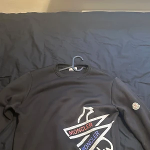 Svart sweatshirt från Moncler - Svart sweatshirt från Moncler med stort tryck i vitt, rött och blått framtill samt klassisk Moncler-logga på ärmen. Tröjan har rund halsringning och långa ärmar. Perfekt för dig som gillar stilrena och exklusiva plagg.