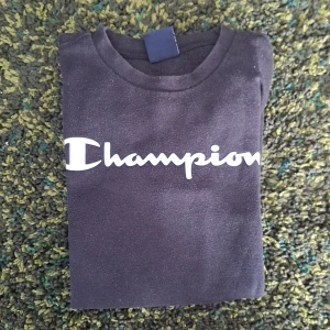 Svart långärmad tröja från Champion - Svart långärmad tröja från Champion med klassisk vit logga framtill. Tröjan har rund halsringning och är tillverkad i mjukt material som passar perfekt till en avslappnad stil. Enkel och stilren design som funkar året runt.