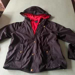 Svart vindjacka från Polo Ralph Lauren - Svart vindjacka med röd insida från Polo Ralph Lauren. Jackan har huva, guldiga knappar och klassisk logga broderad på bröstet. Praktiska fickor framtill och snygg, sportig look. Perfekt för dig som gillar stilrena och funktionella plagg. Pris kan diskuteras😸