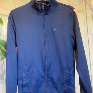 Marinblå trackjacka Tommy Hilfiger - Snygg marinblå trackjacka från Tommy Jeans med dragkedja framtill och logoband längs ärmarna. Koftan har ståkrage, två fickor och diskret broderad logga på bröstet. Perfekt för en sportig och stilren look.