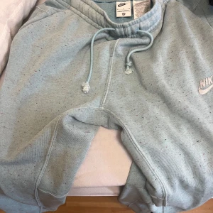 Ljusblå mjukisbyxor från Nike - Säljer ett par ljusblå mjukisbyxor från Nike med diskreta färgstänk och klassisk logga på benet. Byxorna har snörning i midjan och är gjorda i mjuk bomullsmix som är riktigt skön. Perfekt för chill dagar eller när du vill vara extra bekväm.