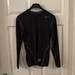 Svart långärmat underställ Nike - Svart långärmad träningströja från Nike Pro Combat med gråa sömmar och diskret Nike-logga på bröstet. Tight passform och tillverkad i syntetmaterial som andas, perfekt för intensiva träningspass eller sport.