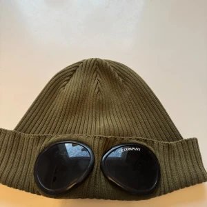 Olivgrön mössa med glasögondetalj från C.P. Company - Snygg olivgrön mössa från C.P. Company med unika glasögondetaljer framtill. Mössan är ribbad och tillverkad i ett mjukt material som ger en skön passform. Perfekt för att ge din outfit en cool touch.