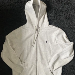 Grå hoodie från Ralph Lauren - Säljer en stilren grå hoodie från Ralph Lauren med dragkedja och klassisk logga på bröstet. Perfekt för en avslappnad look. Den har långa ärmar och en praktisk huva med snörning.
