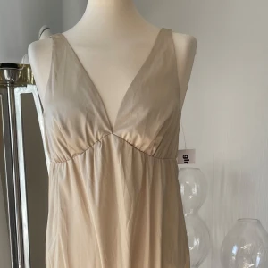Beige klänning från Gina 38 ny  - Snygg beige klänning från Gina med v-ringad design. Klänningen har en elegant och enkel stil, perfekt för olika tillfällen.  Det står topp på kappen men det är en kort klänning 