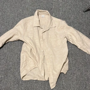 Beige linneskjorta från Lindex 146 - Säljer en stilren beige skjorta från Lindex i storlek 146. Skjortan har långa ärmar och knappar framtill. Perfekt för en avslappnad och snygg look. Tillverkad i ett lätt och bekvämt material.