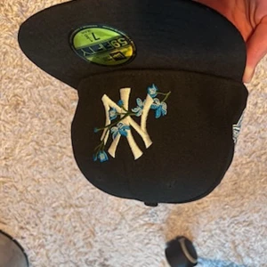 Svart keps från New Era - Snygg svart keps från New Era med broderad logotyp i vitt och blått. Kepsen har en platt skärm och en cool detalj med blommor. Perfekt för att ge din outfit en sportig touch.