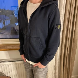 Mörkblå hoodie från Stone Island - Säljer en mörk blå hoodie från Stone Island med dragkedja och huva. Jackan har en broderad logga på ärmen och ribbade muddar. Perfekt för en casual look. STORLEK 14-15 MEN SITTER SOM S
