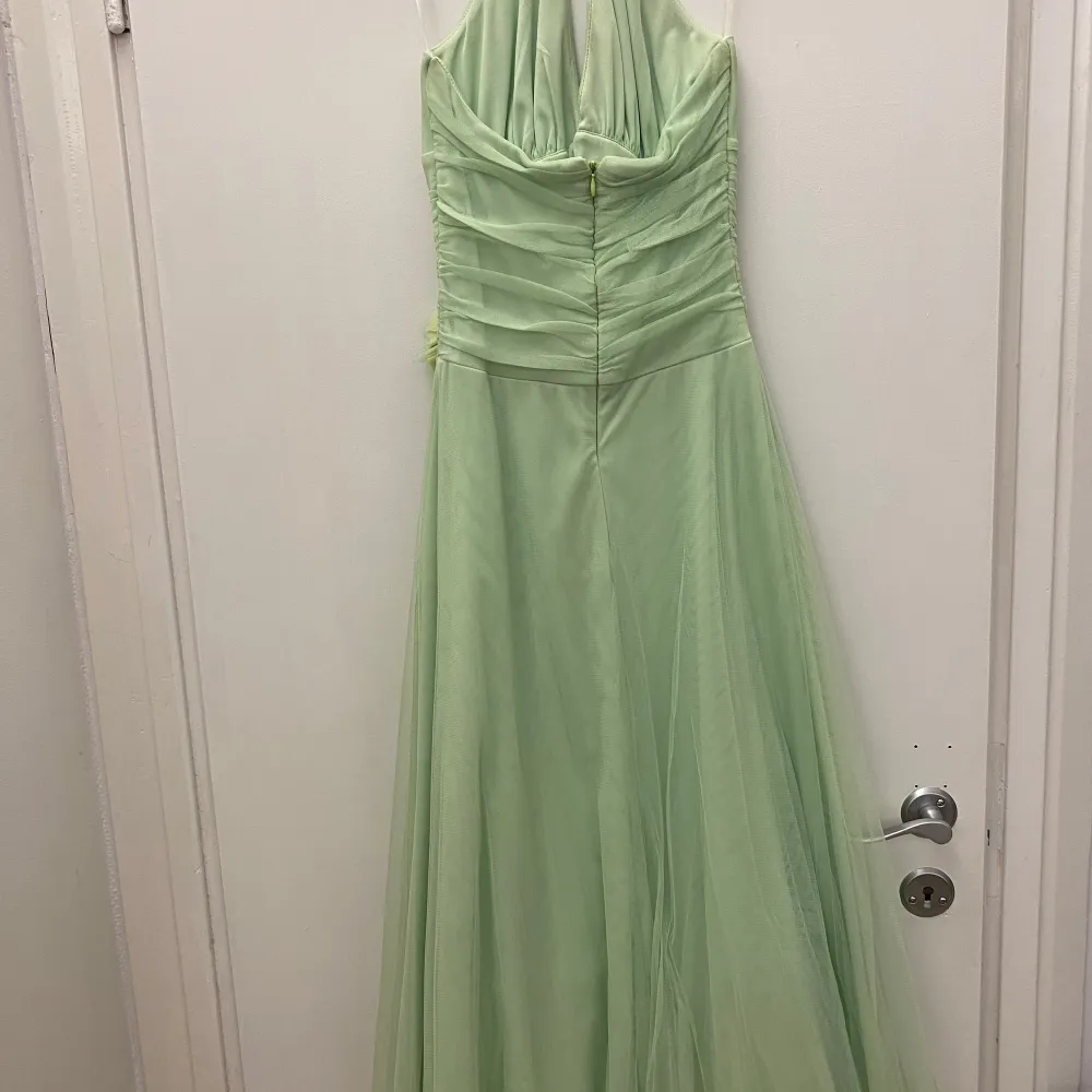 Elegant grön midi-klänning med halterneck och v-ringad design. Klänningen har en vacker blomdetalj i midjan men sim går att flytta. Den är tillverkad i ett lätt och flödande material i tull. Perfekt för speciella tillfällen.. Mekot.
