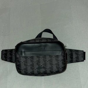 Svart midjeväska från Lacoste - Snygg svart midjeväska från Lacoste med geometriskt mönster och justerbart bälte. Väskan har en dragkedja och insida i kontrasterande orange färg. Perfekt för att bära dina nödvändigheter med stil. Köpt på Zalando. ‼️ENDAST SERIÖSA KÖPARE‼️