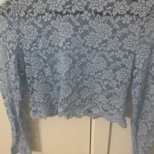 Blå spetsblus - Säljer en elegant blå spetsblus med blommigt mönster. Blusen har långa ärmar och är croppad, vilket ger en modern och stilren look. Perfekt för att lägga till en touch av elegans till din garderob.💗