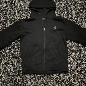 Svart hoodie från Polo Ralph Lauren - Säljer en stilren svart hoodie från Polo Ralph Lauren med dragkedja och klassisk logga på bröstet. Perfekt för en avslappnad stil. Den har en praktisk huva och fickor framtill. Passar bra till både jeans och joggers.